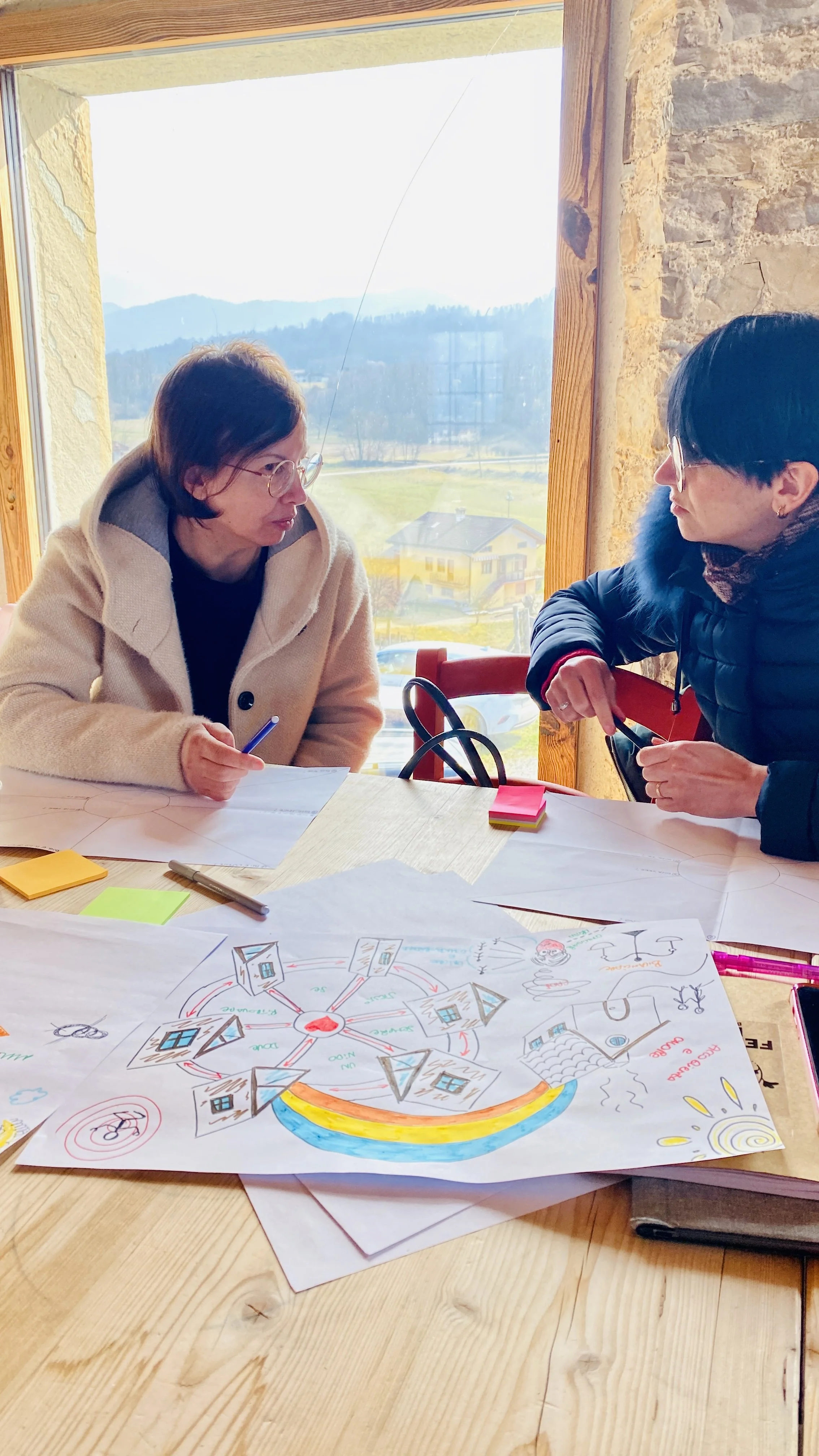 Due donne discutono davanti a un tavolo con disegni e post-it, in una stanza con finestra che mostra paesaggio di montagna.