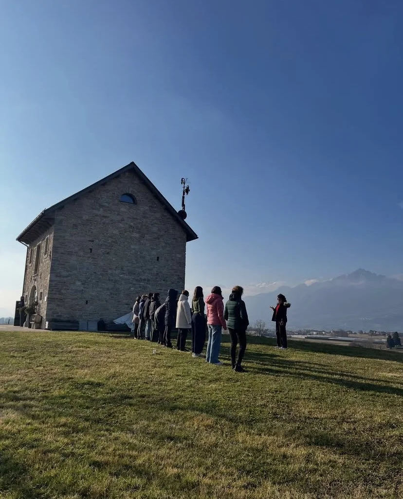 Gruppo di persone in piedi in fila davanti a una piccola chiesa in aperta campagna, con montagne sullo sfondo e un cielo sereno.