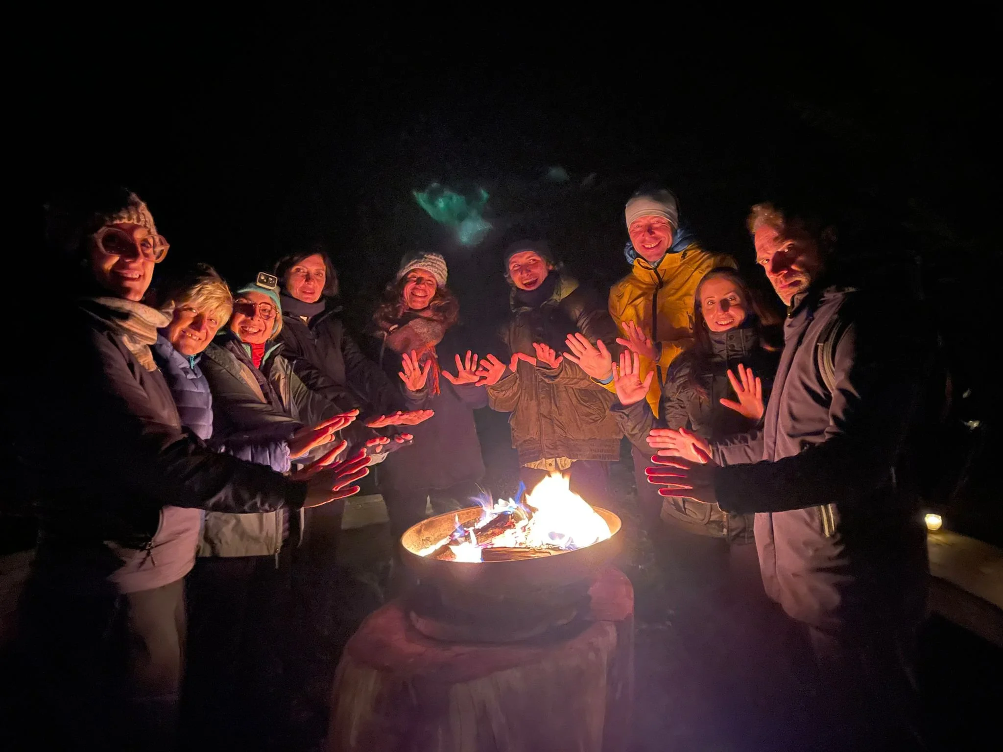 Gruppo di persone in abbigliamento invernale riunite attorno a un falò di notte, tutte sorridenti e con le mani tese verso le fiamme.
