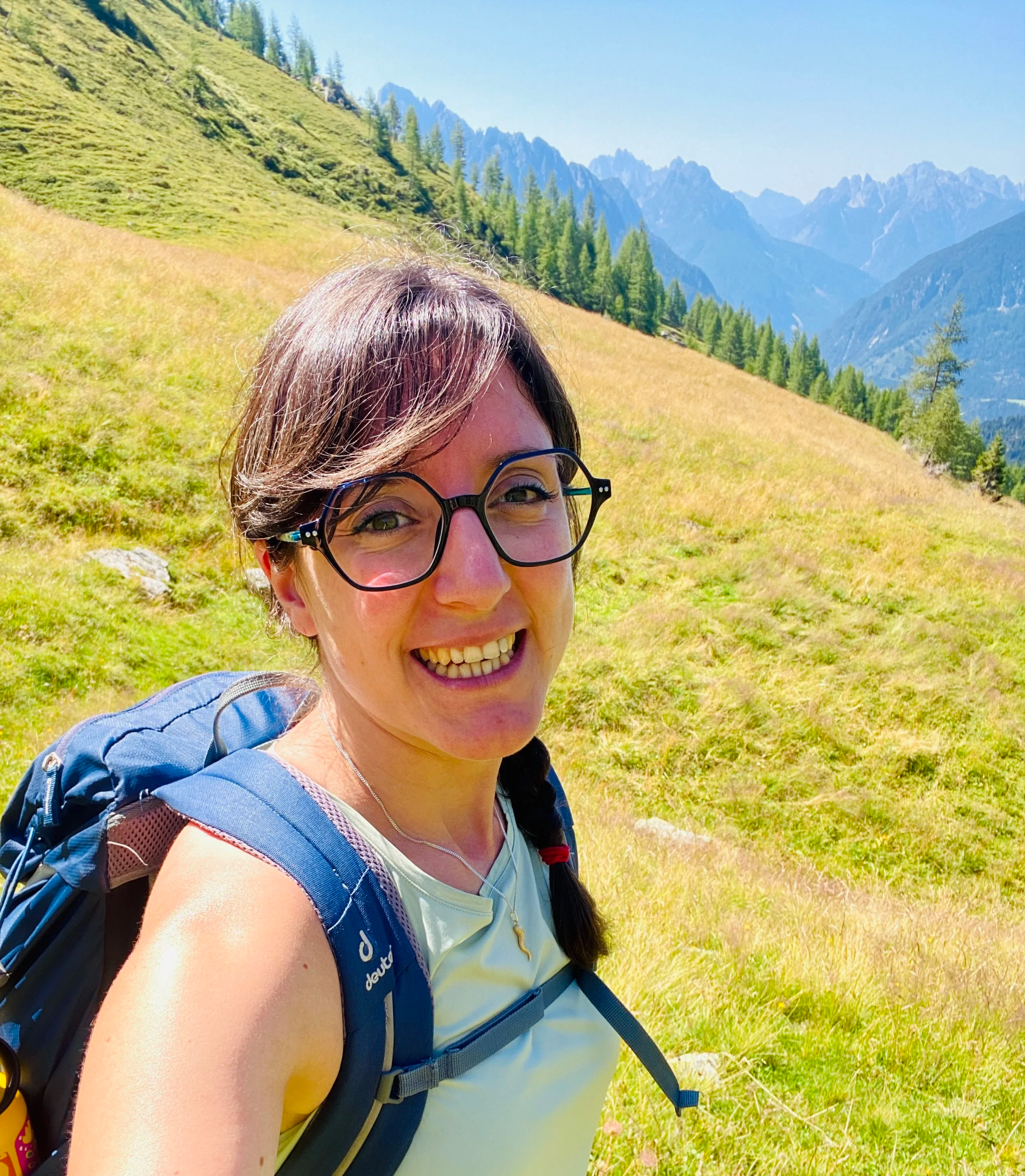 Una donna con occhiali, sorriso e zaino in montagna con paesaggio di colline e montagne verdi e azzurre sullo sfondo.