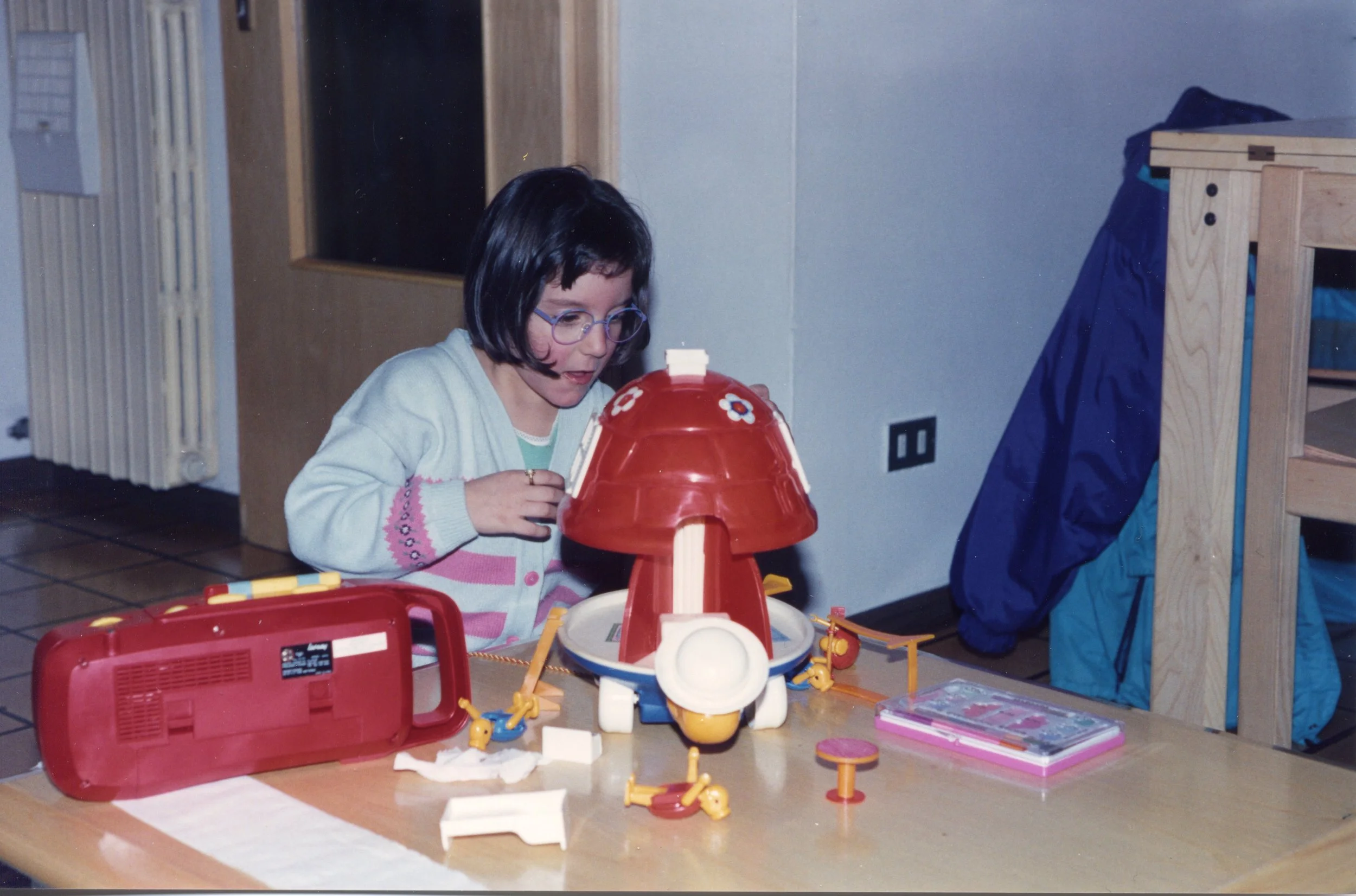 Una bambina con occhiali gioca con la teatrino in plastica rossa a forma di fungo su un tavolo in una stanza.