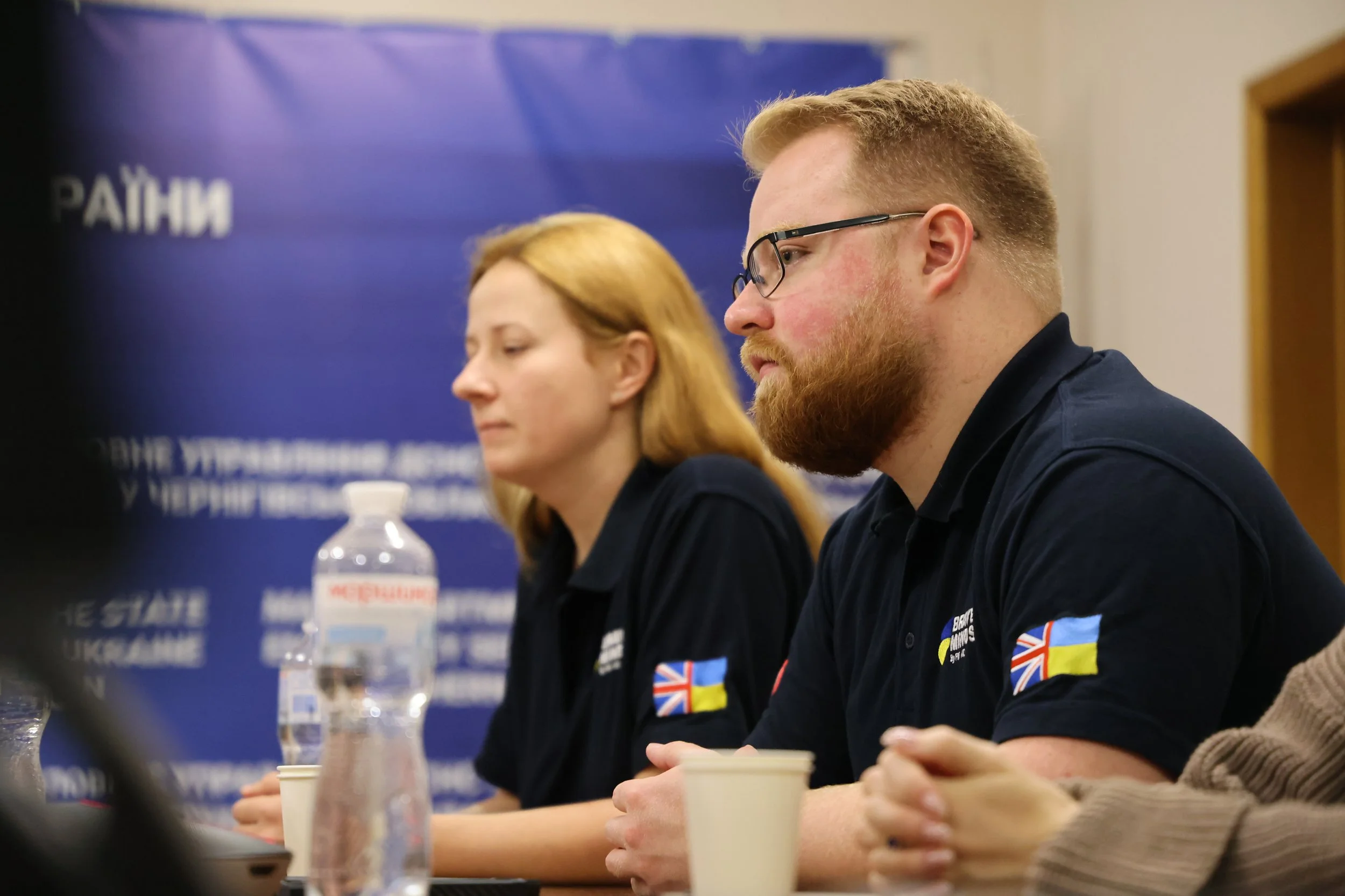 Two people sitting at a table during a press conference or meeting, with water bottles and cups in front of them. The background has a blue banner with some partial text.