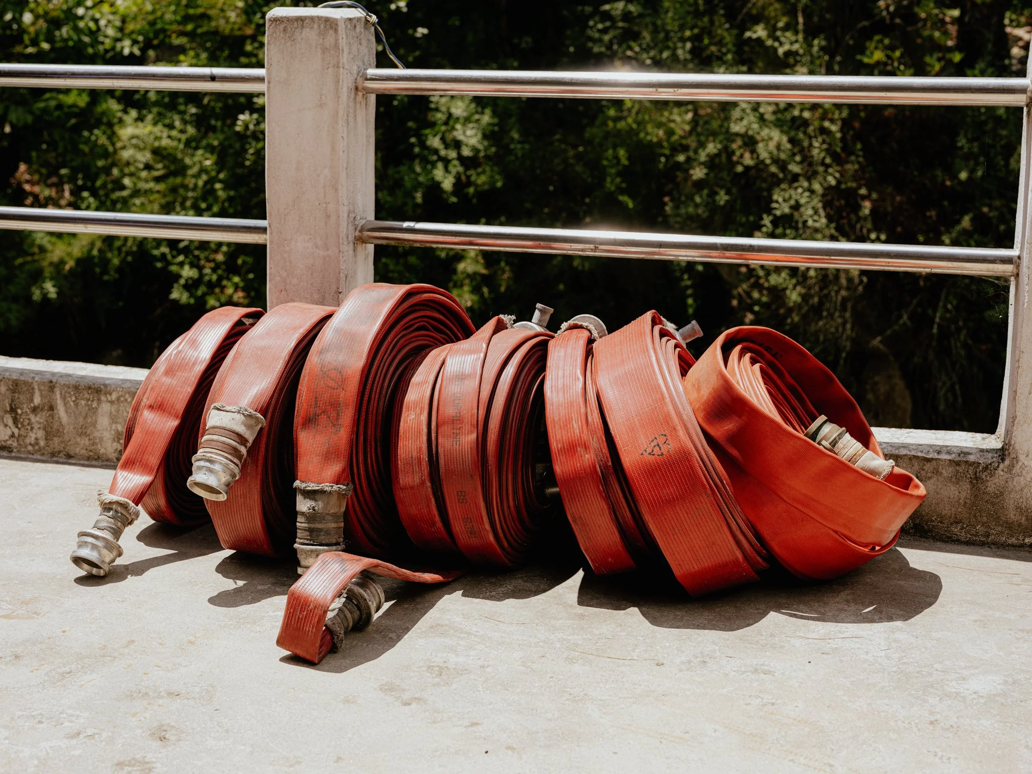 Red fire hoses coiled and placed on a concrete surface, with a metal railing and trees in the background.