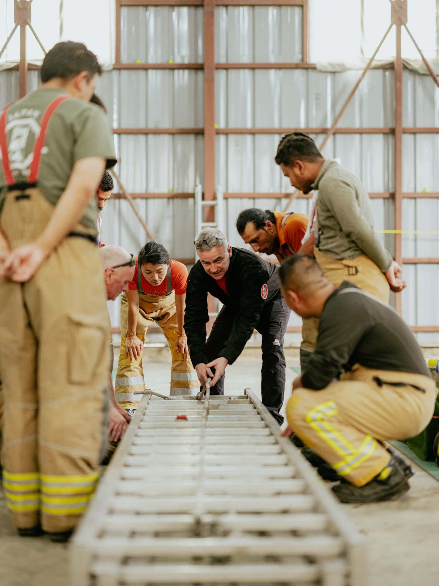Group of people, including firefighters, gathered around and working on a ladder in a warehouse or large garage.