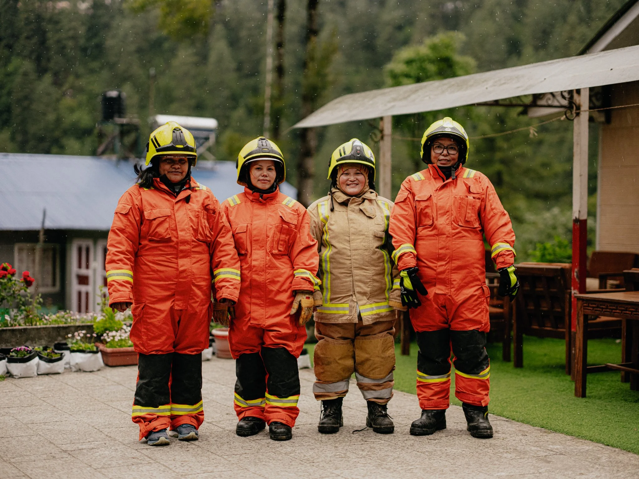 Four firefighters in orange and tan protective firefighting gear standing in a row, with a green, wooded outdoor background, on a paved area.