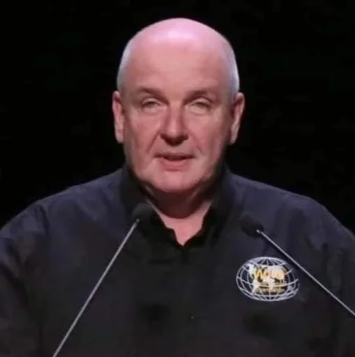 Bald man speaking at a podium, wearing a black shirt with a logo that includes the NASA worm logo and a globe.