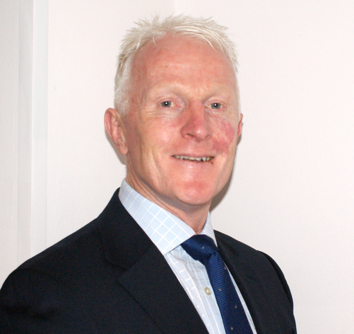 A middle-aged man with white hair in a suit and tie, smiling, against a plain white wall background.