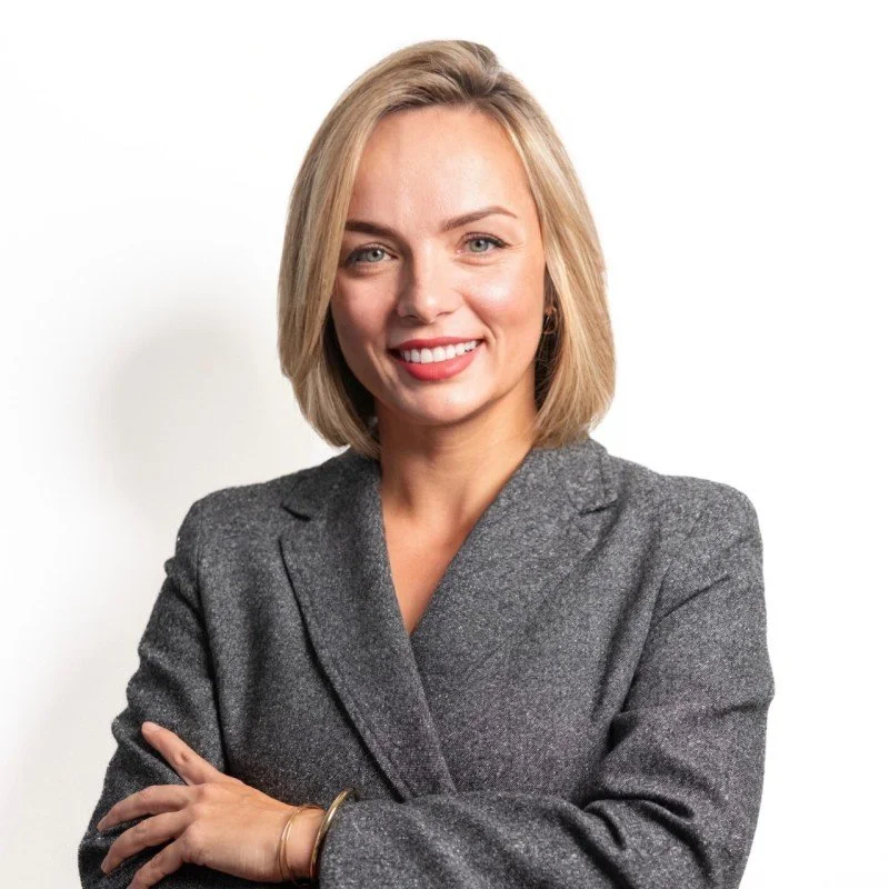 A woman with blonde hair and a professional gray blazer, smiling with arms crossed.