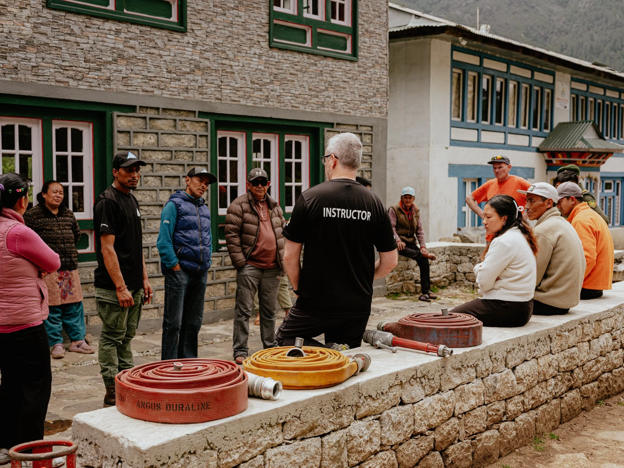 Group of people listening to a man wearing an 'Instructor' shirt during a training session outdoors, with coiled hoses on a stone ledge in front of a building with colorful window frames.