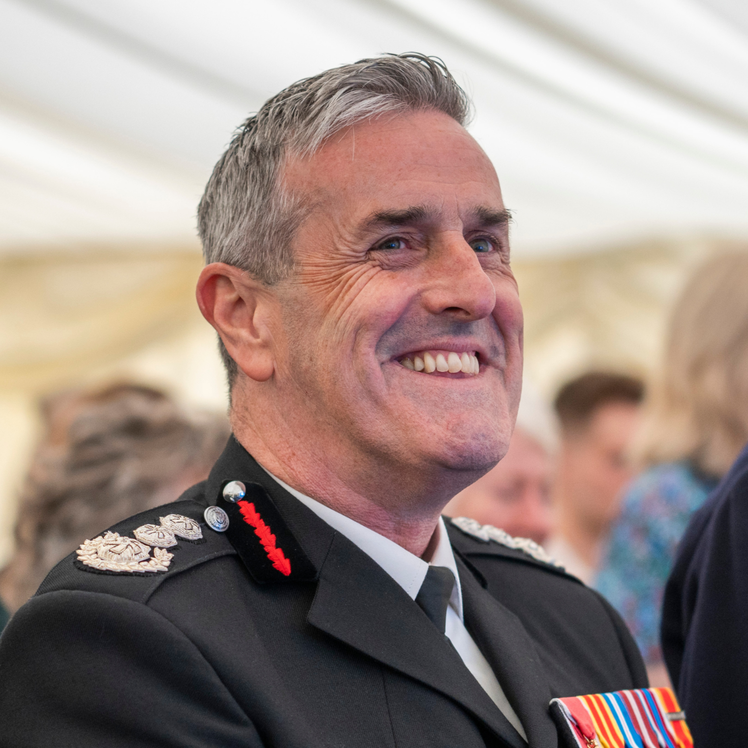 A smiling man in military uniform with medals on his chest, short gray hair, standing in a decorated indoor setting.