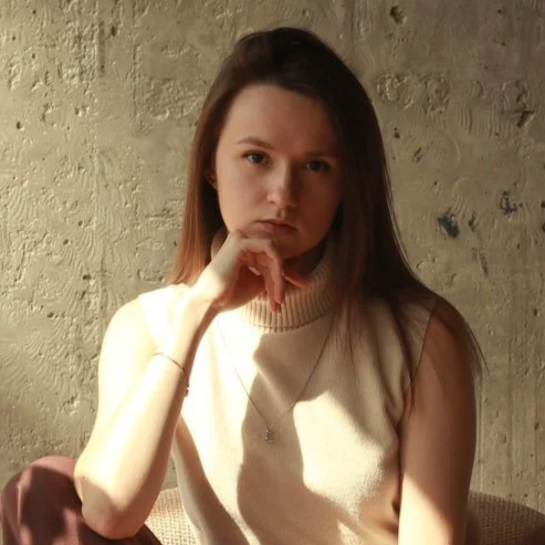 Young woman with long brown hair wearing a sleeveless beige top, sitting against a textured wall, resting her chin on her hand, looking serious.