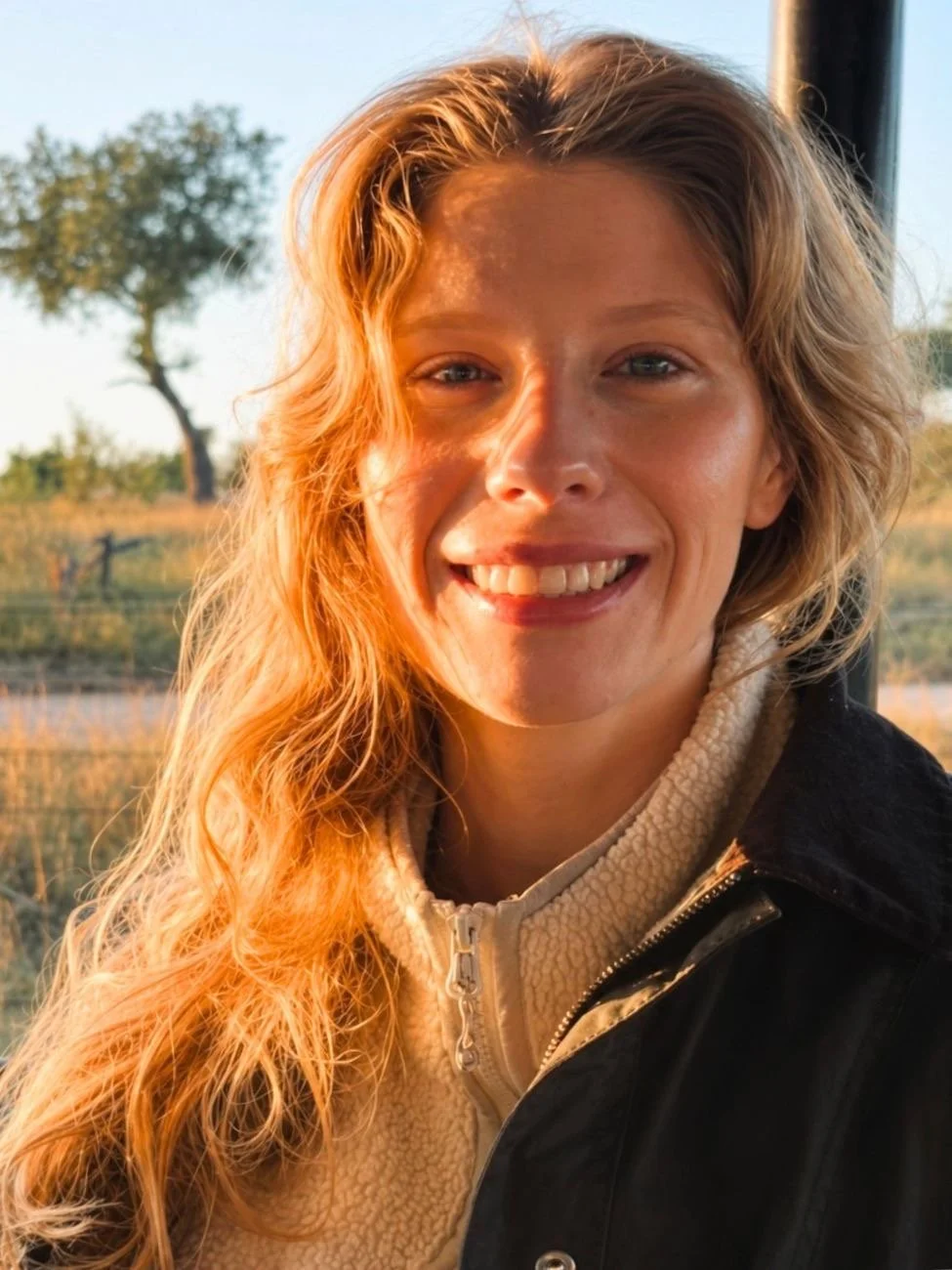 A woman with long wavy blonde hair smiling outdoors during sunset, wearing a beige fleece jacket and a black outer jacket.
