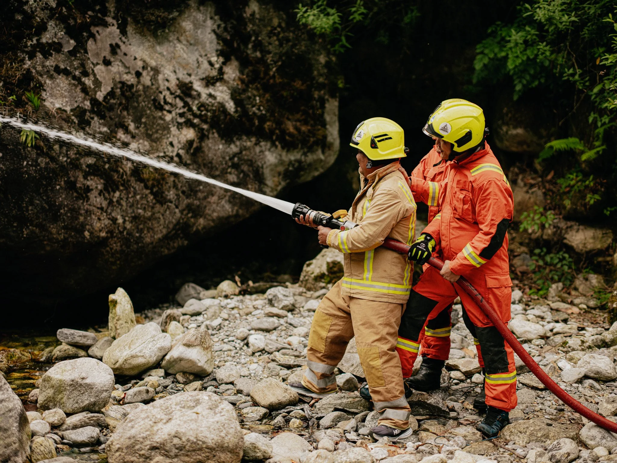 Three firefighters in protective gear spraying water into a rocky, wooded area.