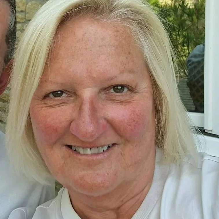 Close-up of a smiling woman with blonde hair and freckles, standing outdoors near a window.