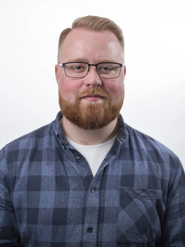 Portrait of a man with glasses, red hair, and a beard wearing a blue plaid shirt against a white background.