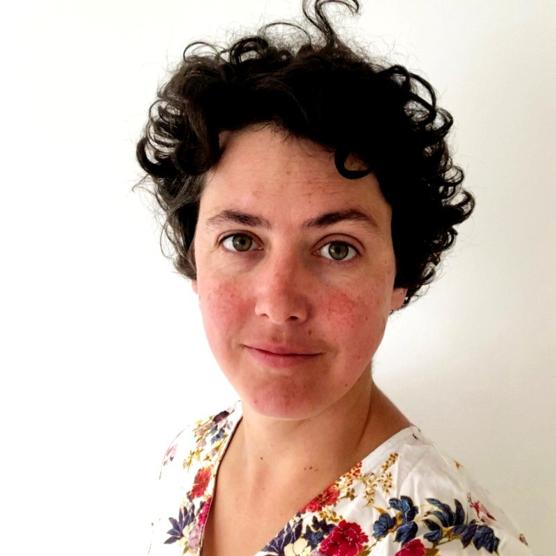A woman with short, curly dark hair and light skin taking a selfie against a plain white wall. She is wearing a colorful floral top and has a slight smile.