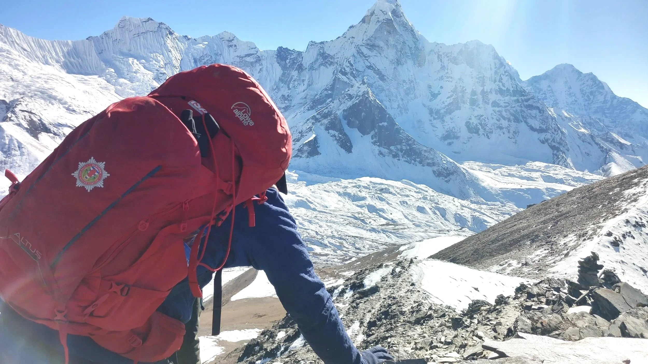 A climber with a red backpack near snow-covered mountains under a clear blue sky.