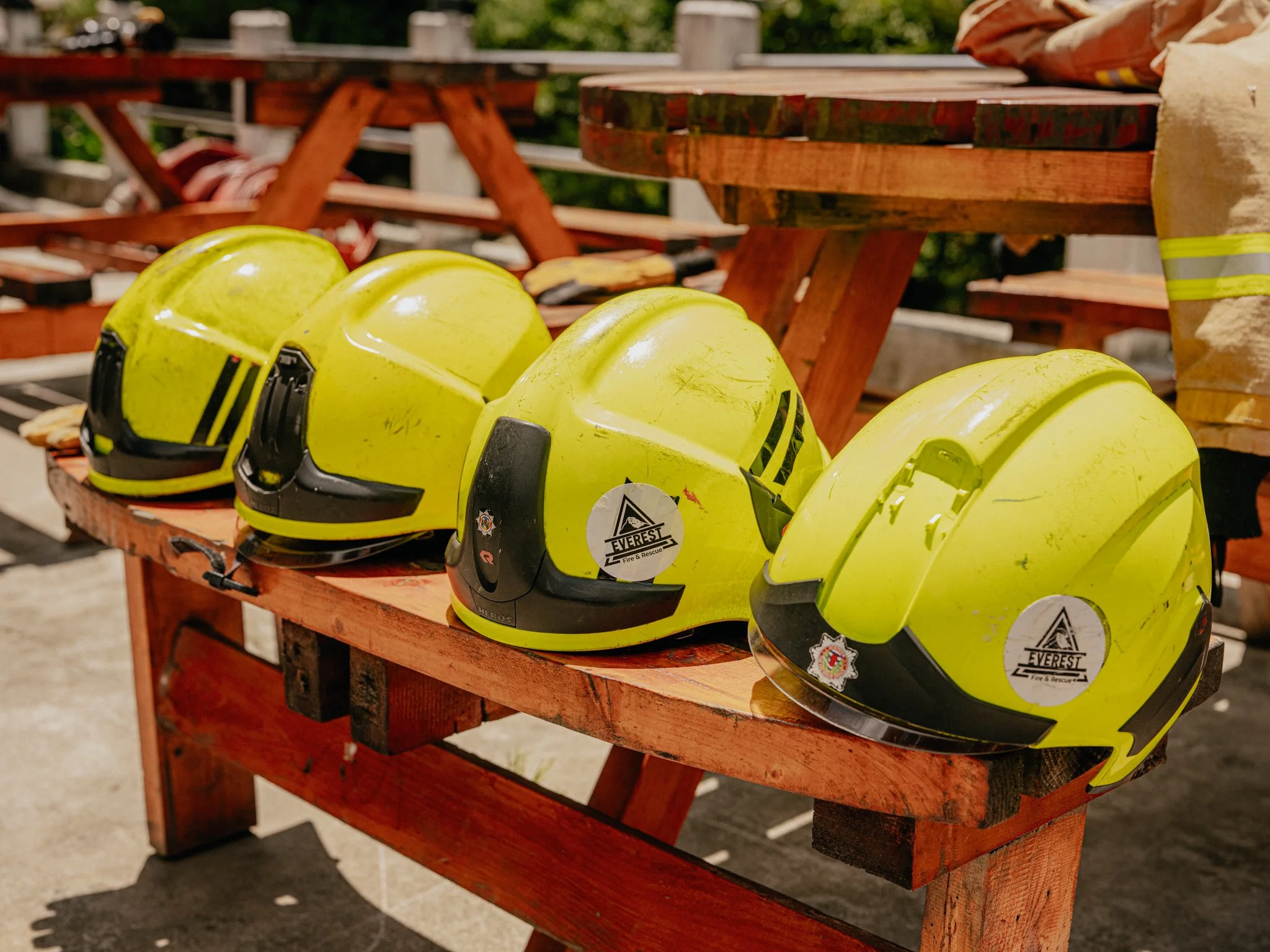 A row of yellow firefighter helmets on a wooden bench outdoors, with other firefighting equipment and trees in the background.