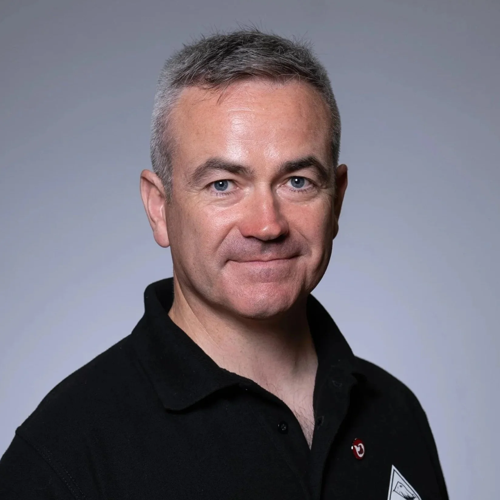 Close-up portrait of a man with short gray hair and blue eyes, wearing a black collared shirt, against a plain light gray background.