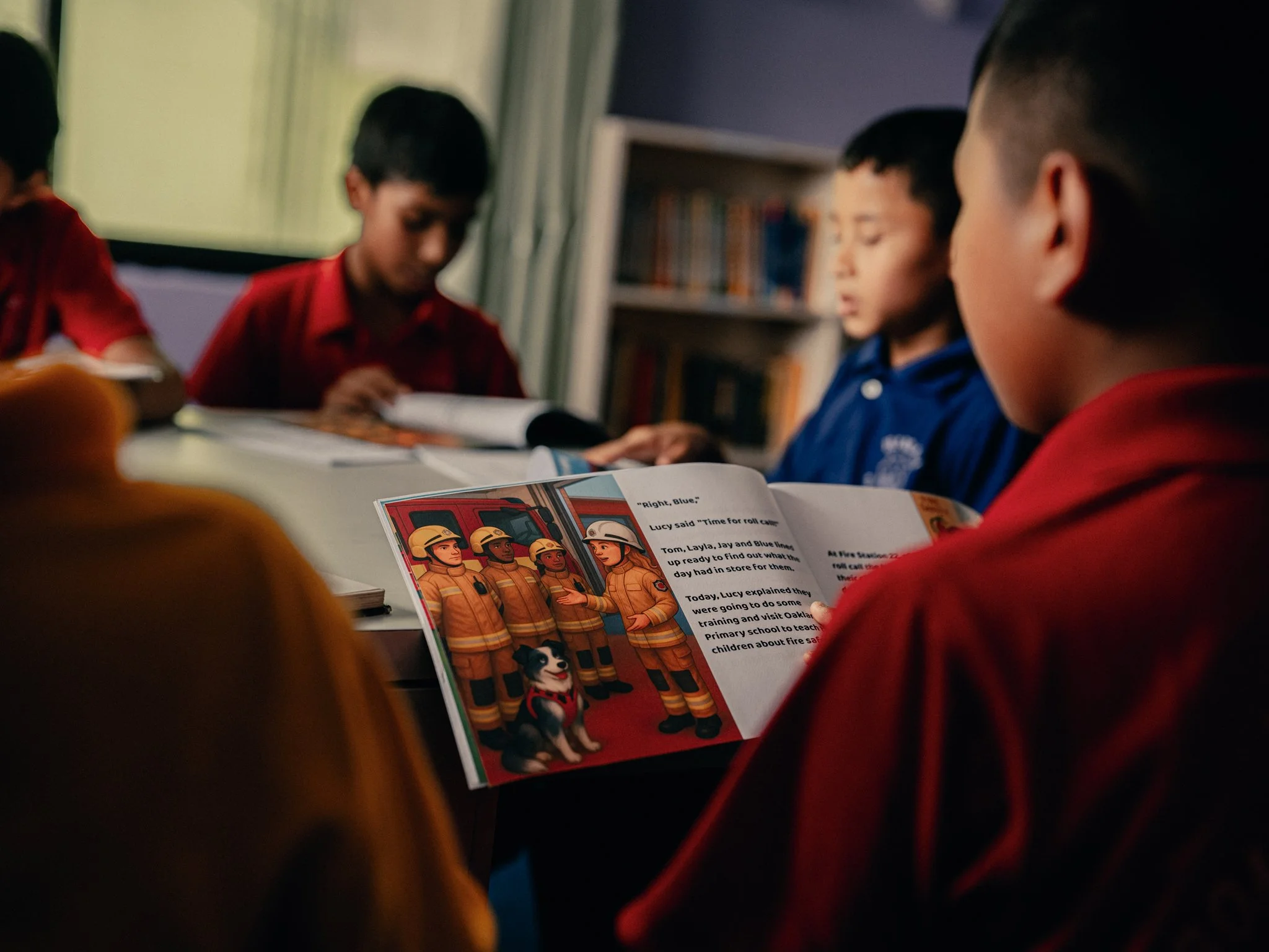 A group of children sitting around a table reading a picture book about firefighters, with the focus on the open page showing cartoon firefighters and text.