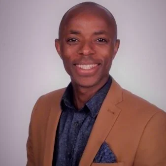 A man smiling, wearing a tan blazer over a dark blue shirt, with a plain light background.