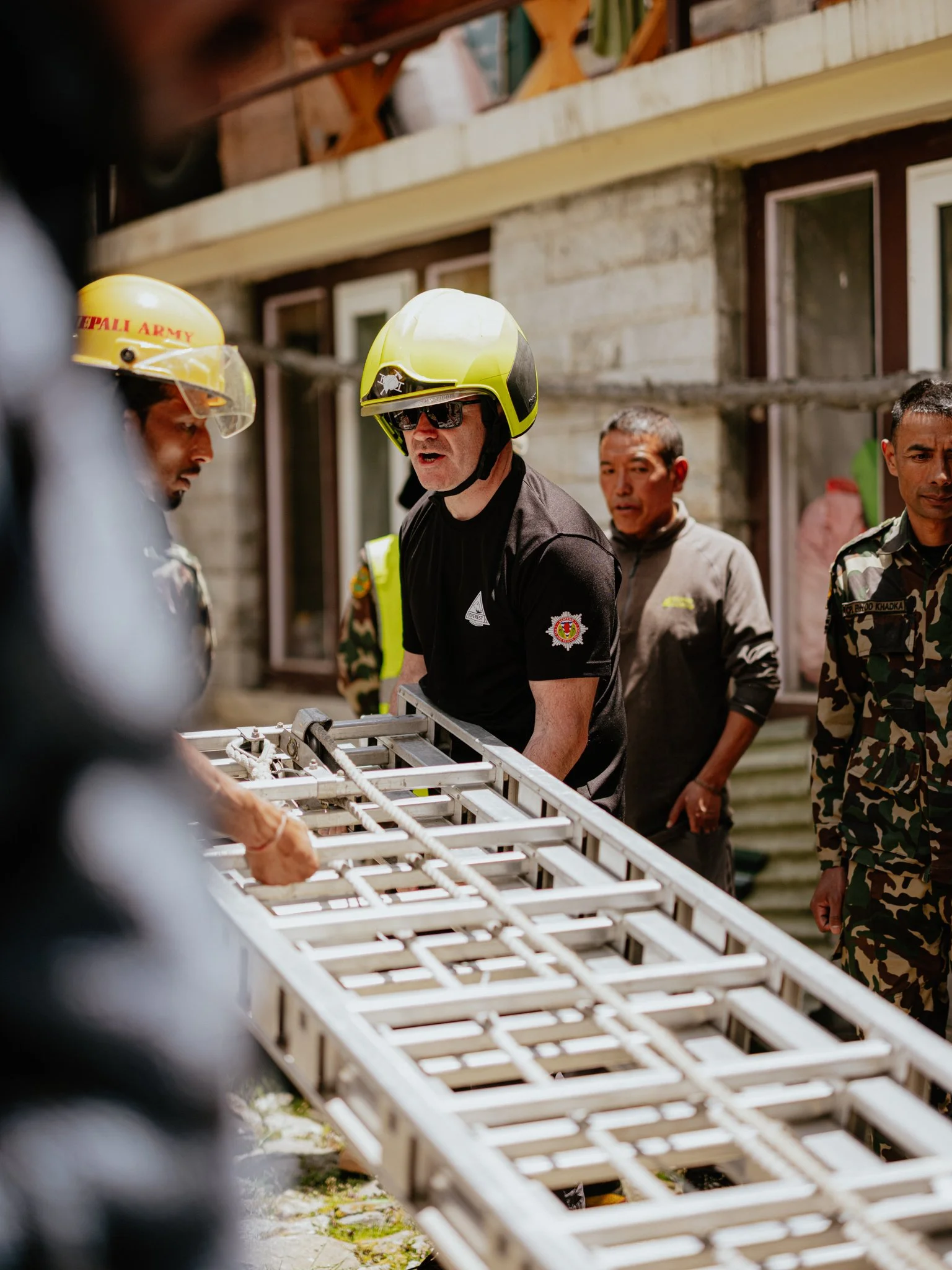 Group of individuals, including firefighters wearing helmets and military personnel, working together outside near a metal structure.