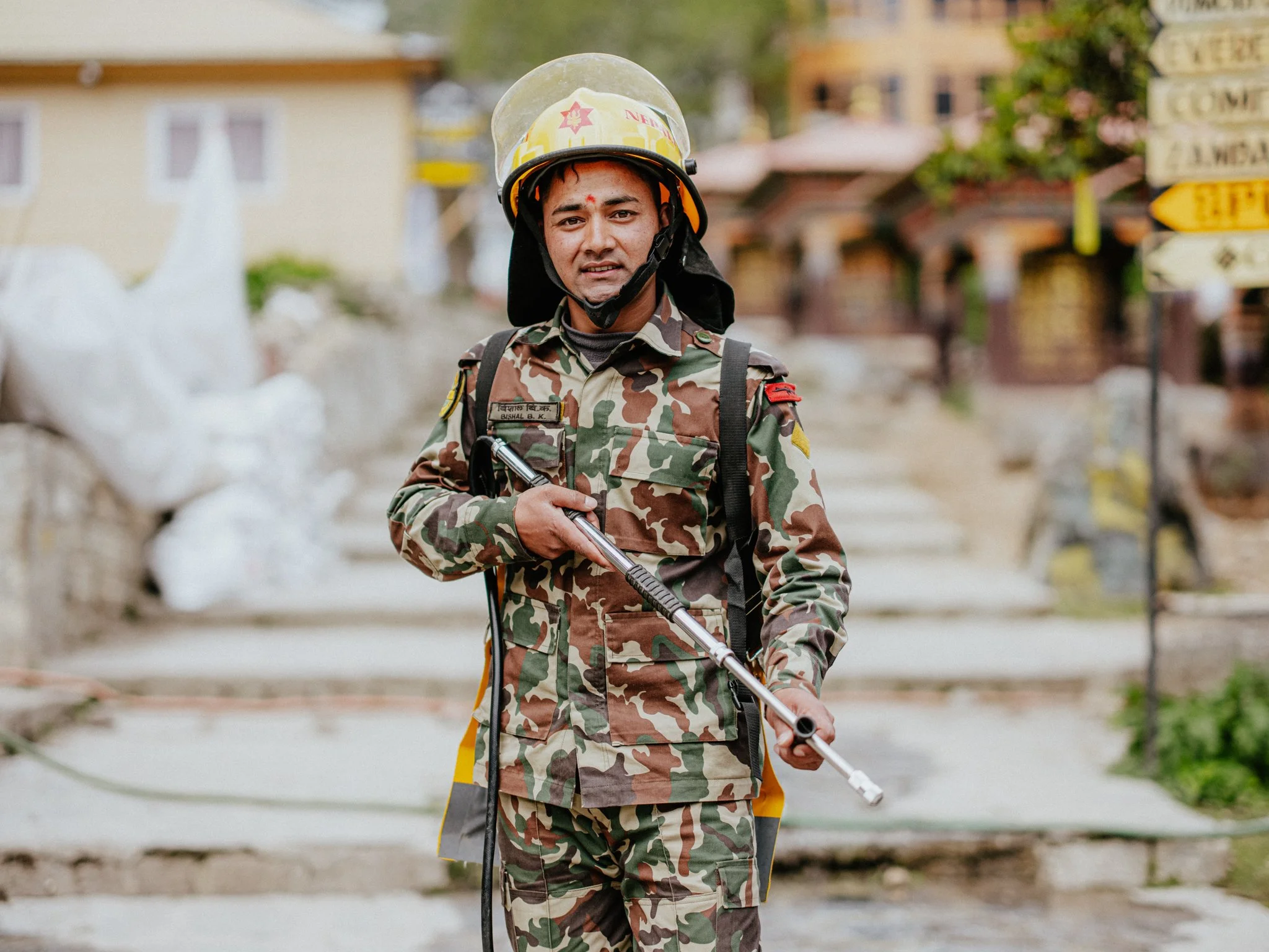 A man wearing camouflage firefighter uniform and a yellow helmet with a face shield, holding a tool, standing outdoors.
