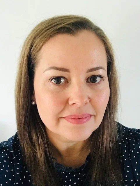 Close-up of a woman with shoulder-length brown hair, wearing a dark blue top with white polka dots, looking at the camera with a neutral expression.