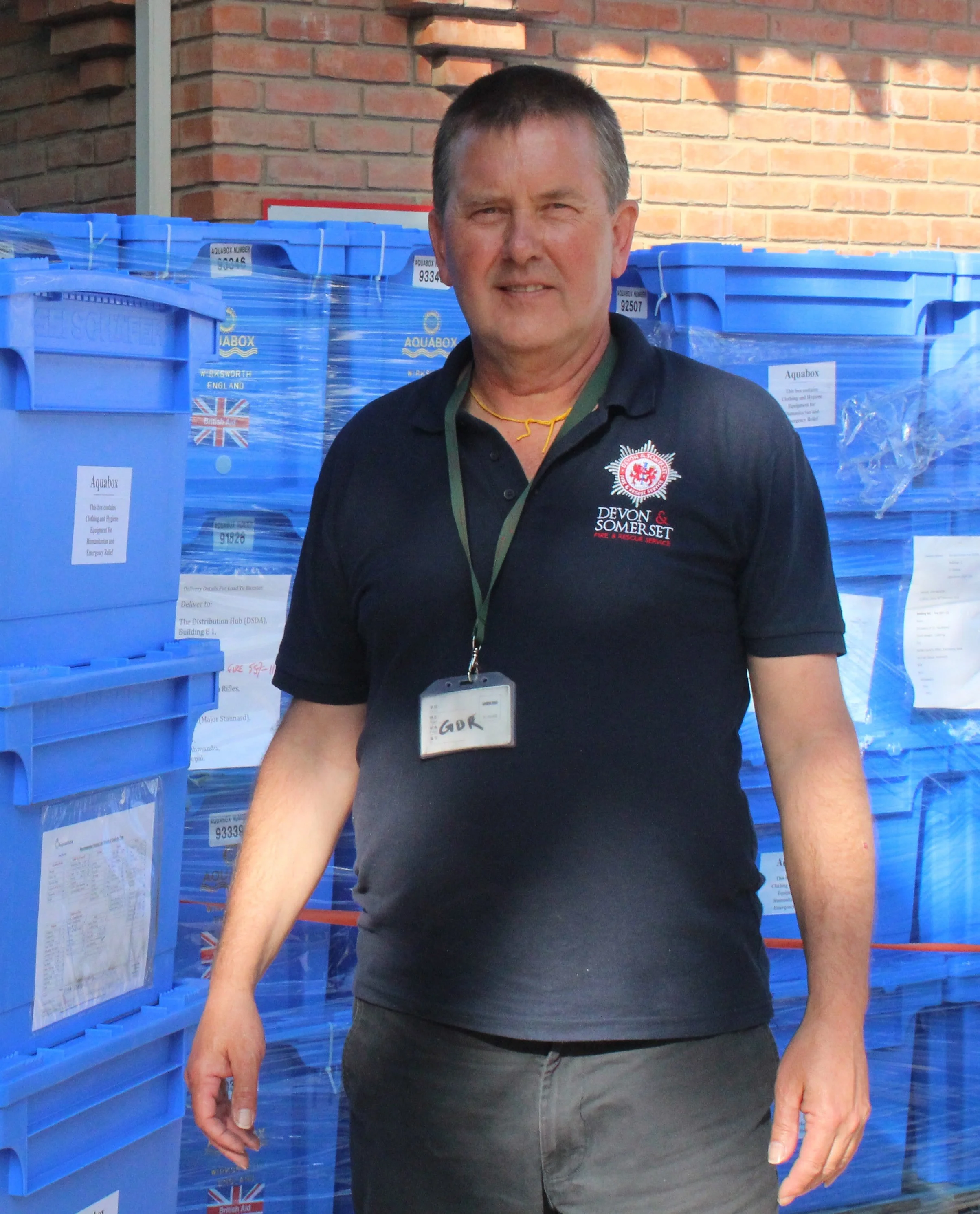 A man standing outdoors in front of stacks of blue Aquabox containers, wearing a navy polo shirt with a Devon & Somerset Fire & Rescue Service emblem and a lanyard with an ID badge. The background shows a brick wall.
