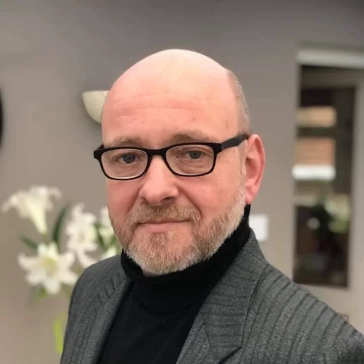 Man met brillen, baard en een pak, staand binnen naast bloemen.