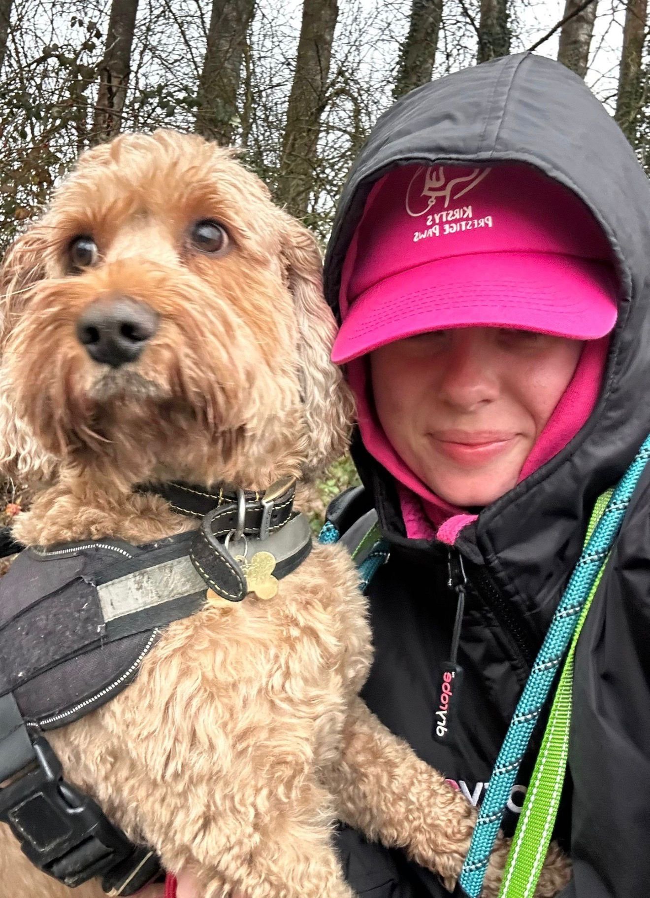 A woman dressed in outdoor gear holding a light brown, curly-haired dog with a harness, in a wooded area during daytime.