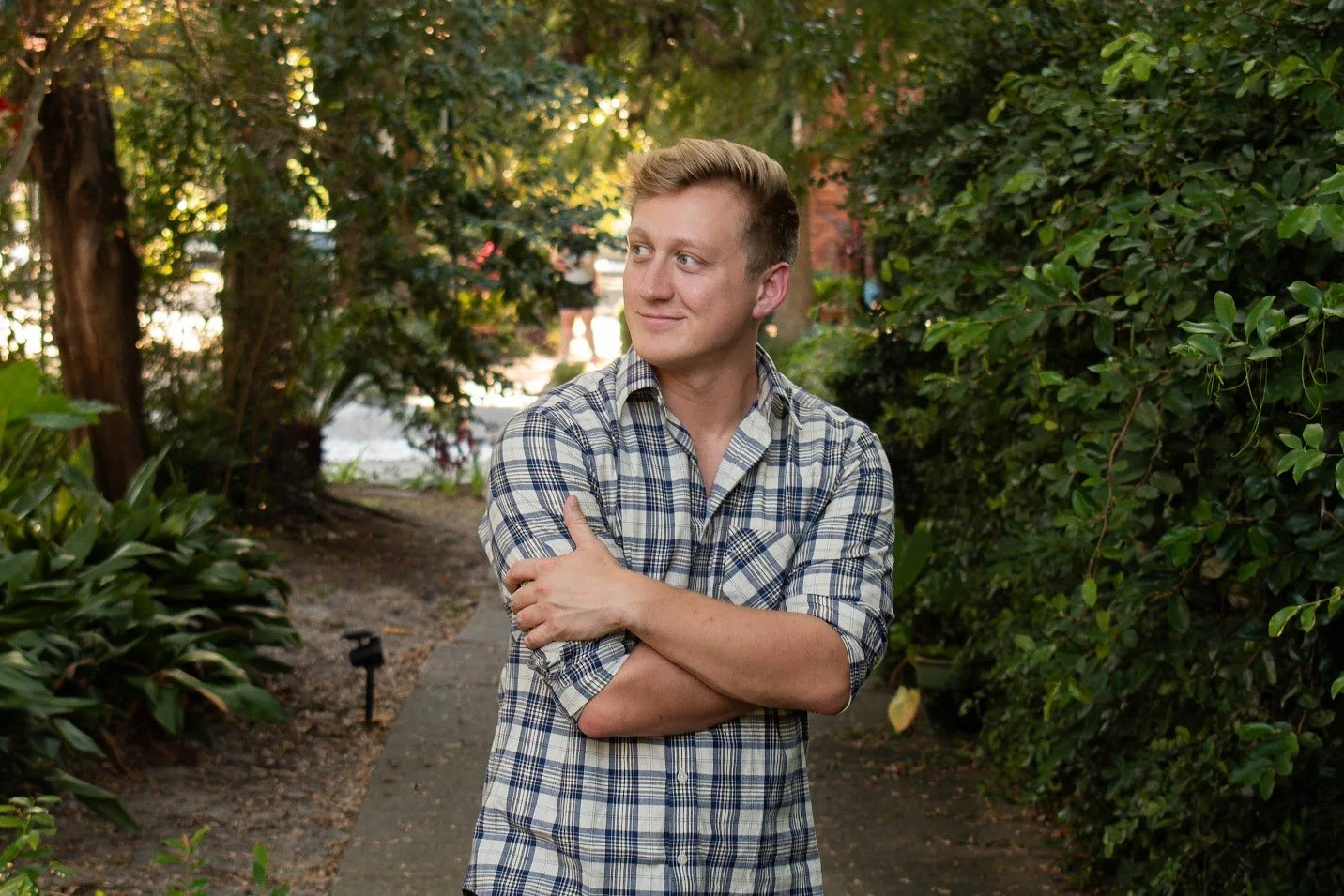 A young man with blonde hair and a plaid shirt crossing his arms, standing on a garden path surrounded by greenery and trees.