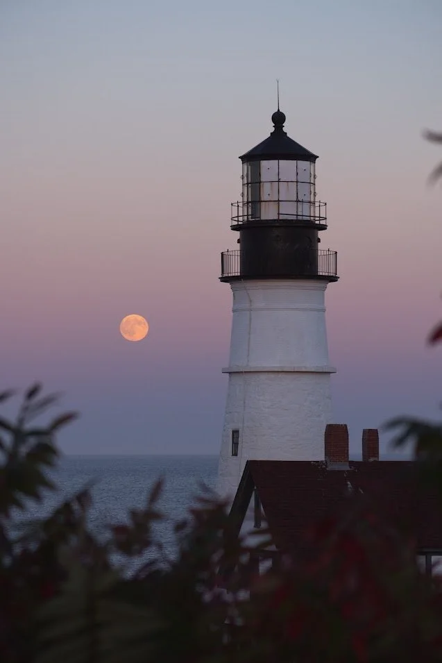 A lighthouse on the coast during dusk, with the moon visible in the cloudy sky.