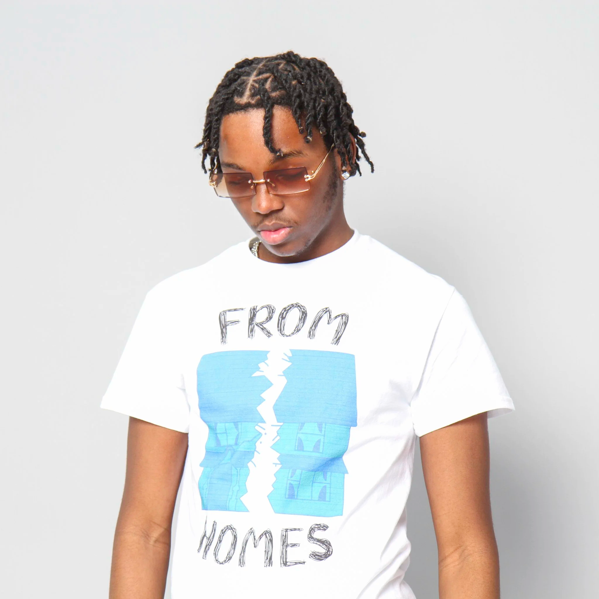 A young man wearing sunglasses and a white T-shirt with a blue graphic and the words 'From Homes' printed on it, standing against a plain light gray background.