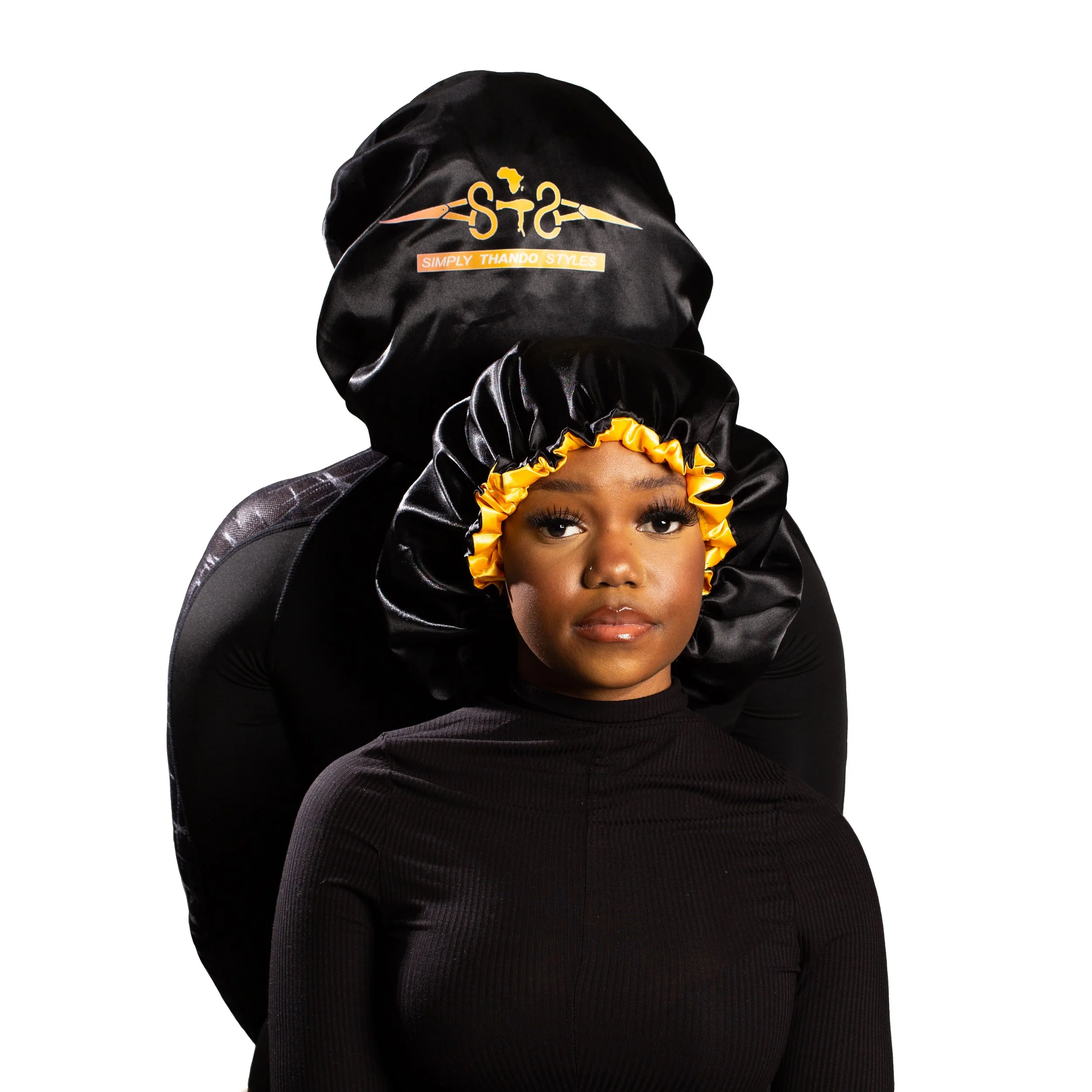A young woman wearing a satin bonnet with yellow and black ruffled trim, standing in front of a large black head wrap with a logo, against a white background.
