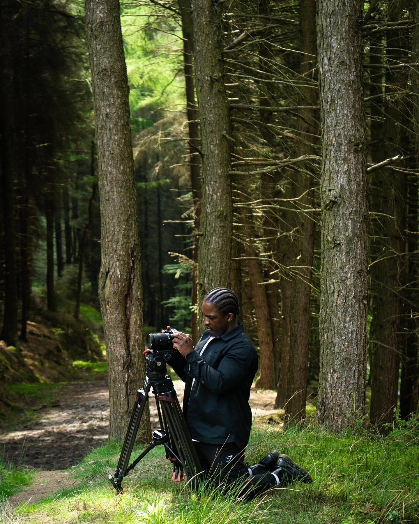 A person with braided hair kneels on the grass in a forest, adjusting a camera on a tripod.