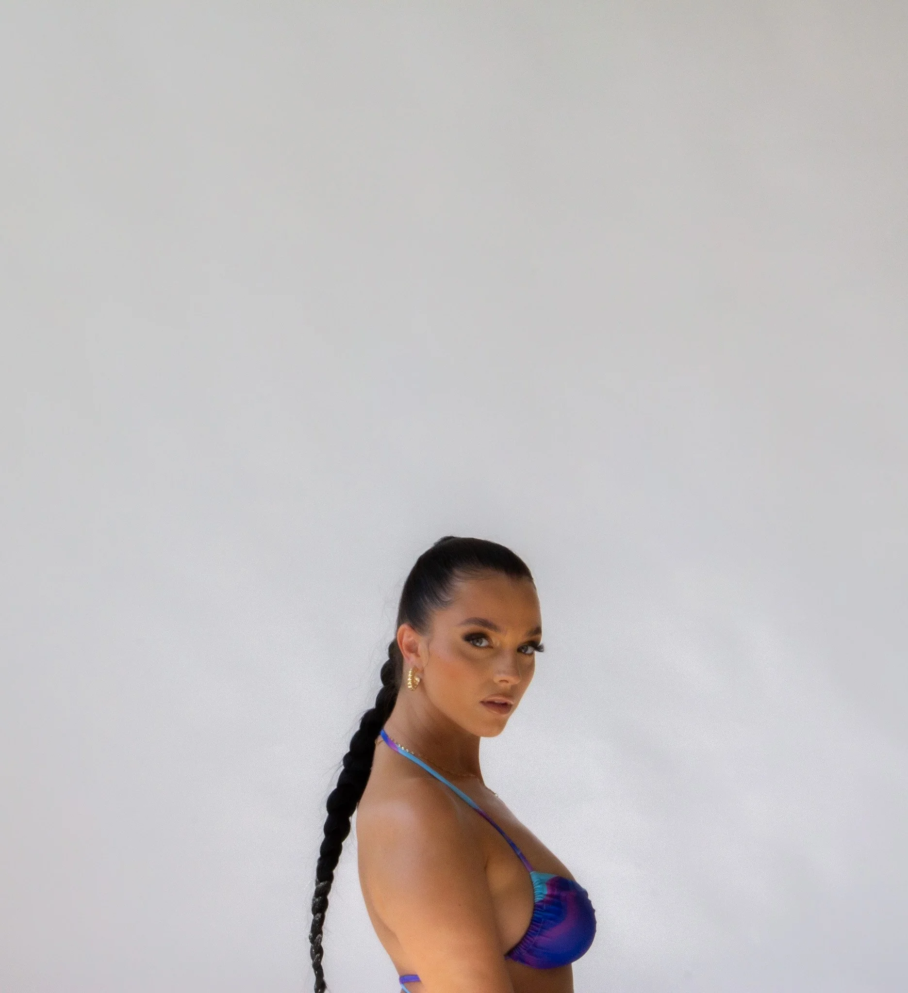 A woman with long braided hair and gold earrings wearing a colorful bikini topless with blue and purple hues, standing against a plain white background.