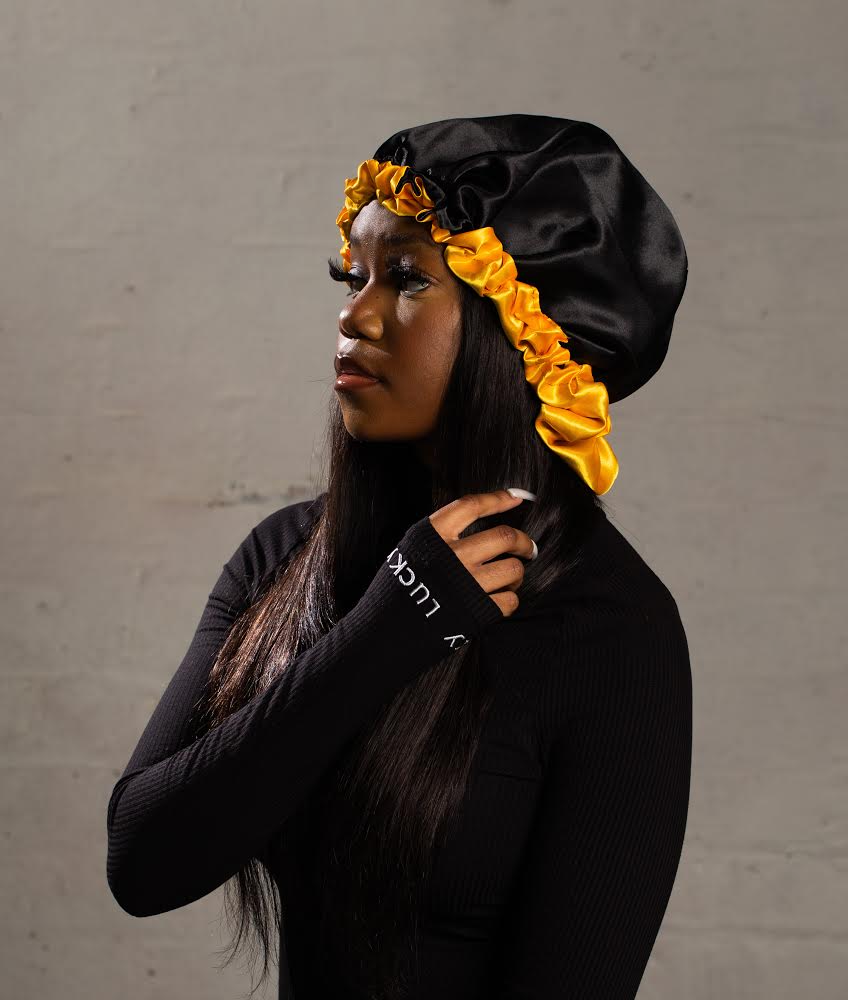 A woman wearing a black satin bonnet with yellow ruffled trim, a black long-sleeve shirt, and long dark hair, standing against a plain gray background.