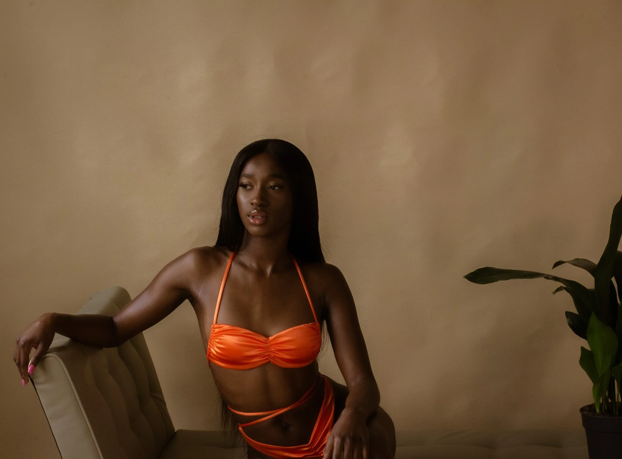 A woman with dark skin and long black hair wearing an orange bikini, sitting on a beige chair against a plain tan wall with a potted plant to her right.