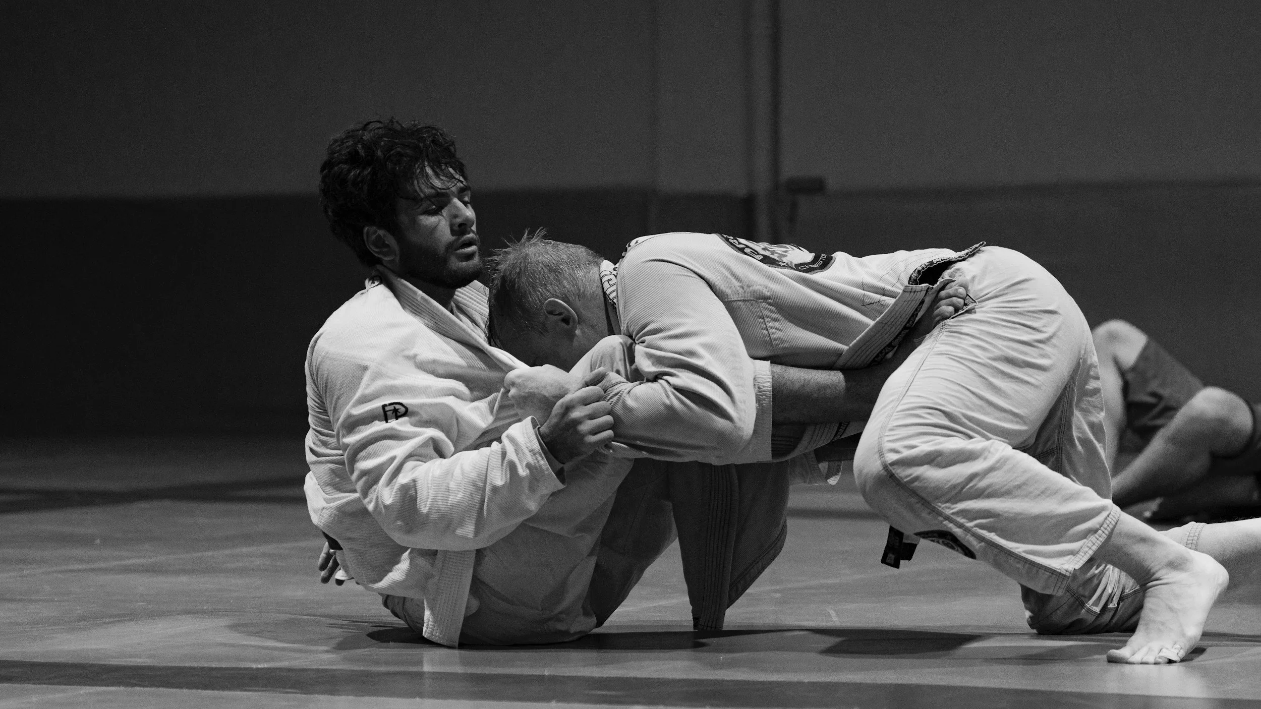 Two male jiu-jitsu practitioners in gis fighting on the mat, one on top and the other on the bottom, in a competitive match.