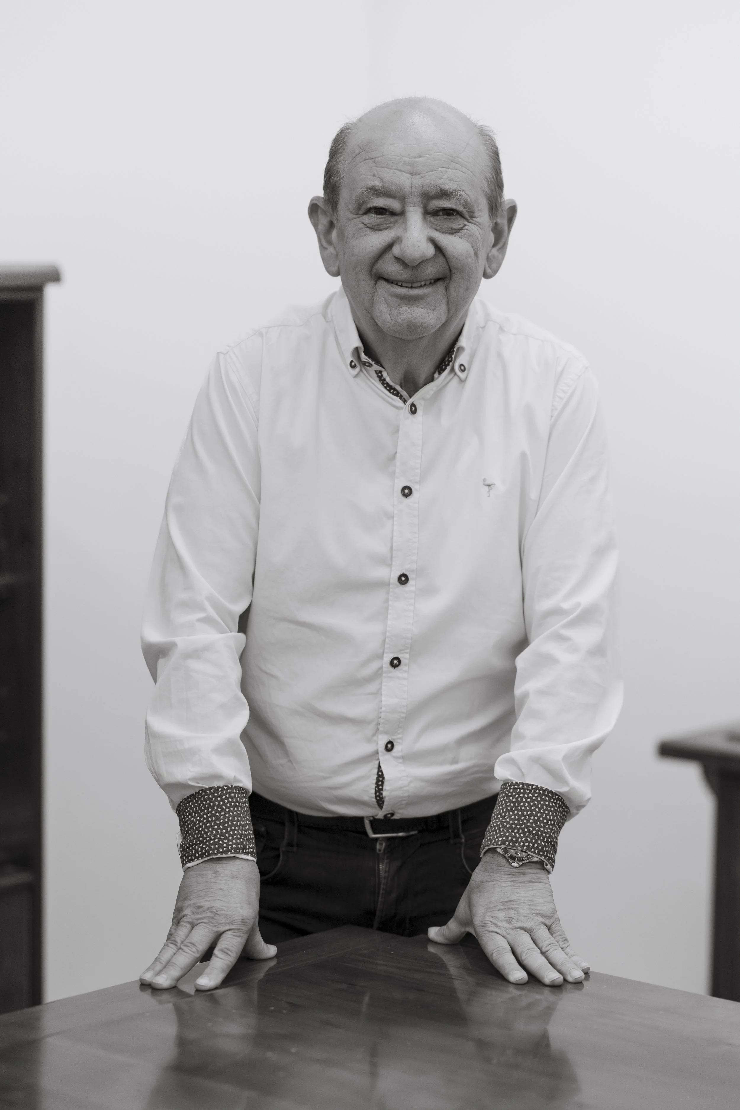 Hombre mayor con camisa blanca y joyas, apoyado en una mesa de madera, sonriente, en un ambiente de oficina.