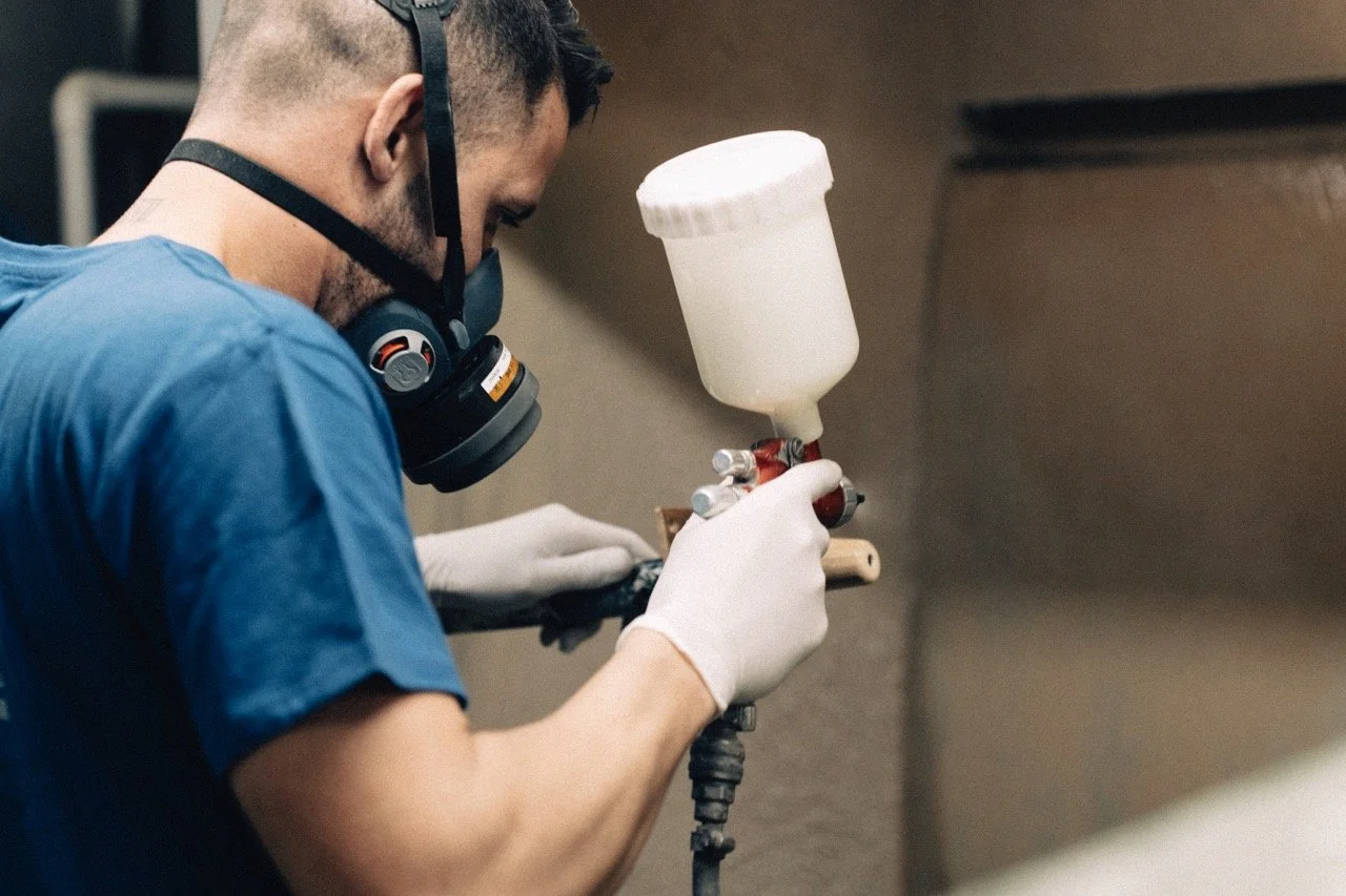 Persona usando máscara y guantes, trabajando con una pistola de pintura en un ambiente cerrado.