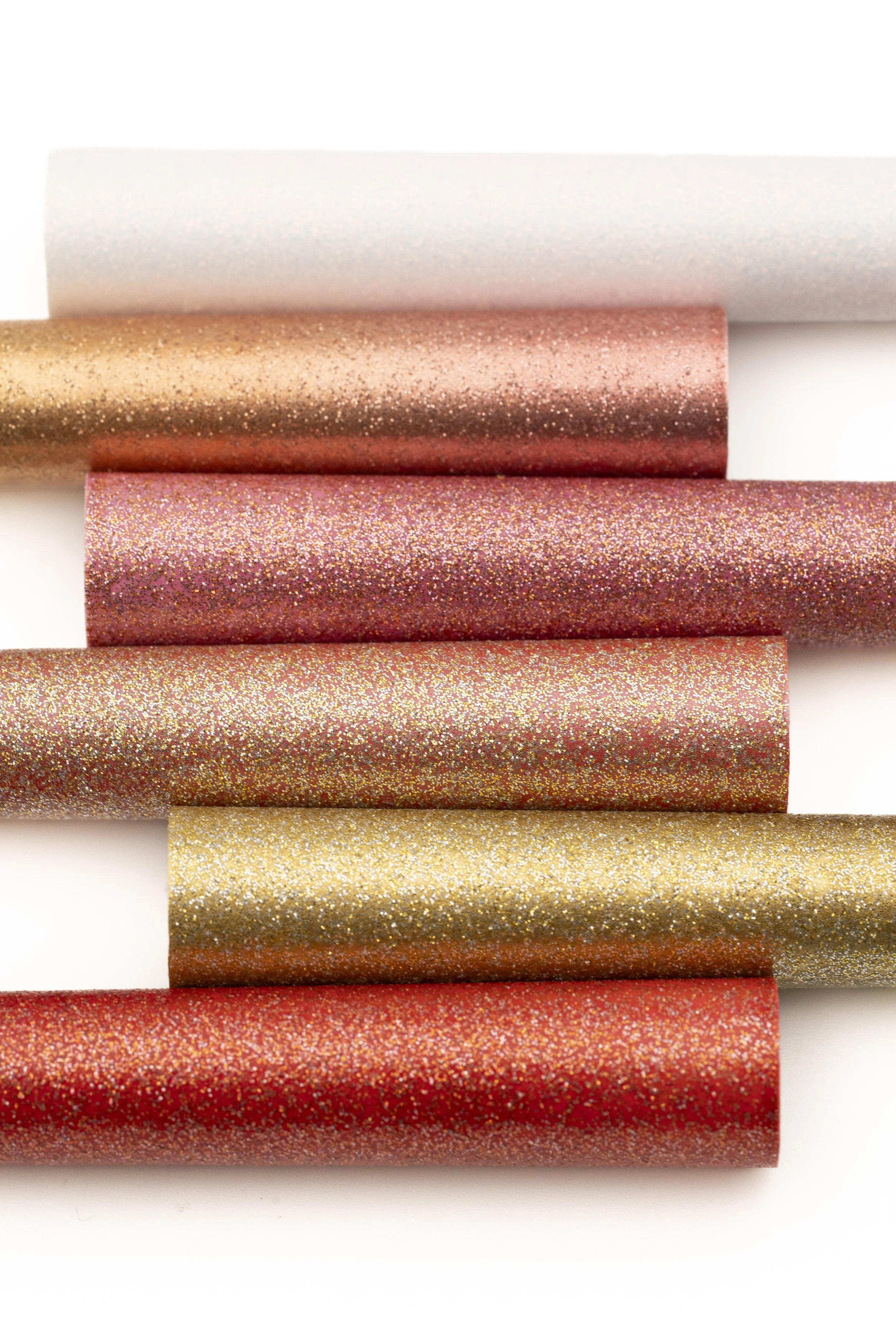 Tiras de papel decorativo con acabado brillante y glitter en tonos dorados, rosados, rojos y dorados, colocadas en fila horizontalmente sobre fondo blanco.
