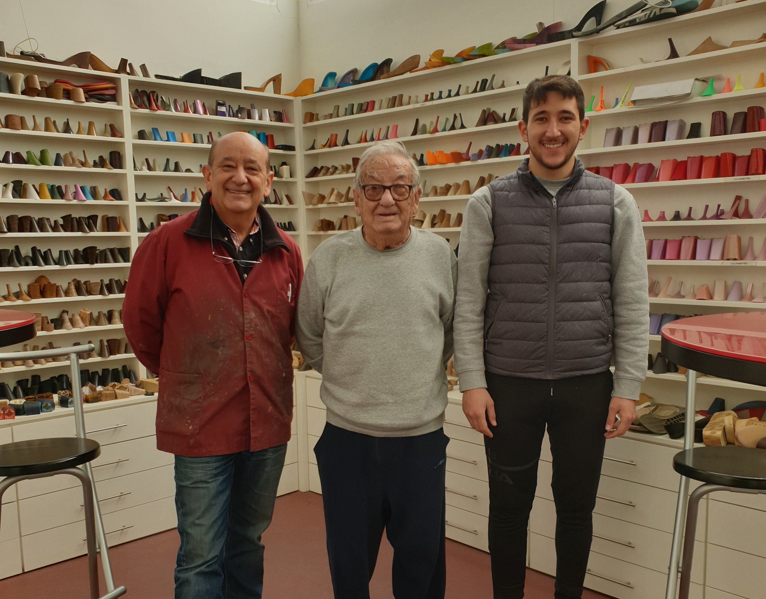 Tres personas de pie en una tienda de zapatos, con estanterías llenas de sandalias y zapatos en el fondo.