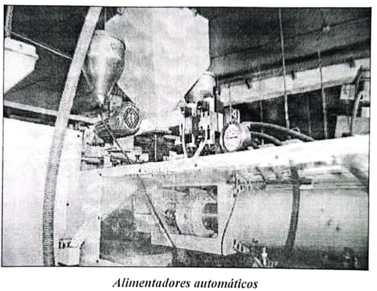 Imagen en blanco y negro de alimentadores automáticos en una fábrica o taller, con maquinaria y tubos visibles.