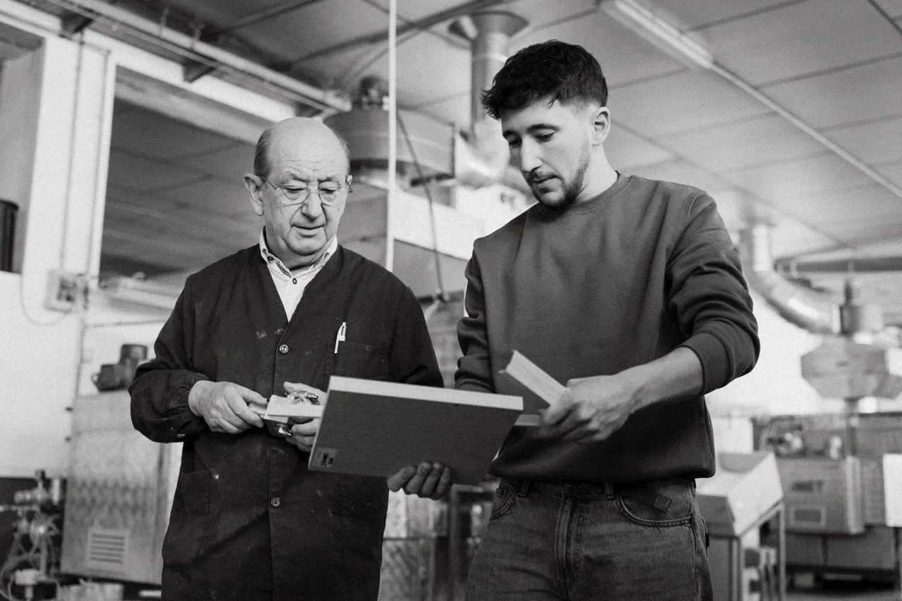 Dos hombres en una fábrica, uno mayor y uno más joven, revisando documentos o instrucciones.