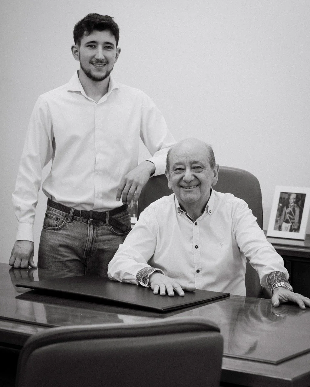 Dos hombres en una oficina, uno mayor sentado en una mesa y otro más joven de pie a su lado, ambos sonriendo, en una fotografía en blanco y negro.