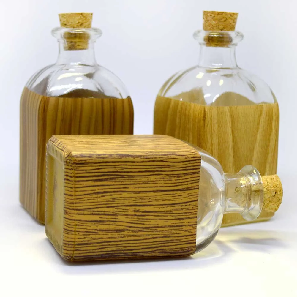 Tres frascos de vidrio con tapas de corcho, decorados con envolturas de papel con apariencia de madera, uno de ellos recostado frente a los otros dos