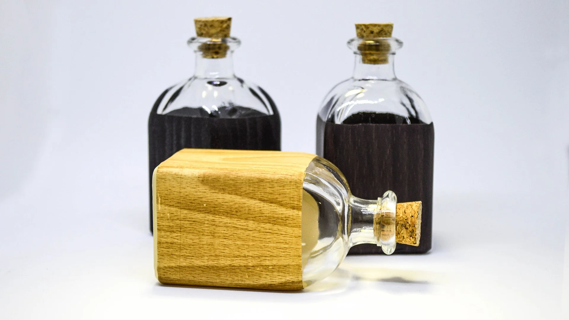 Frascos de vidrio con líquido oscuro, sellados con corcho, uno tumbado con una base de madera, dos de pie detrás en un fondo blanco.