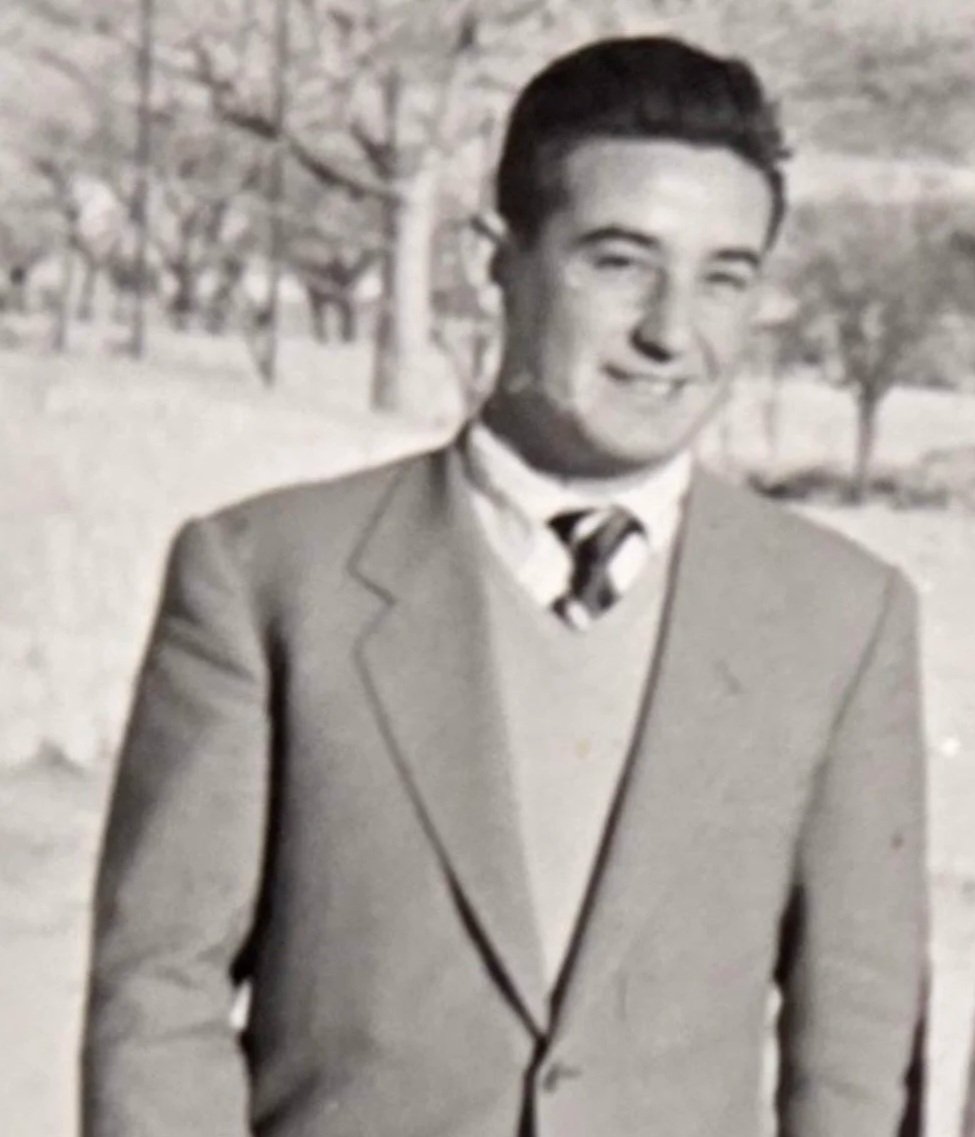 Joven sonriendo en un exterior con árboles en el fondo, vestido con saco y corbata.
